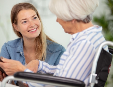 a young woman kneels beside a senior woman's wheelchair to speak with her