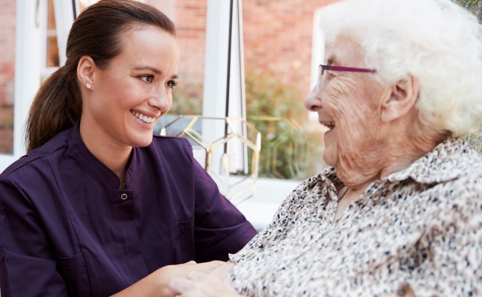 a smiling dietition kneels down next to a seated senior woman for a conversation